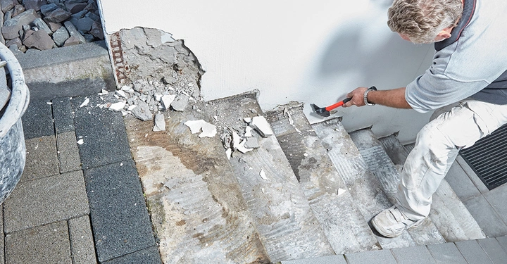 Ein Handwerker bei der Untergrundvorbereitung einer Außentreppe. Mit Hammer und Meißel werden alte Fliesen und Reste von Fliesenkleber für eine saubere Oberfläche entfernt.