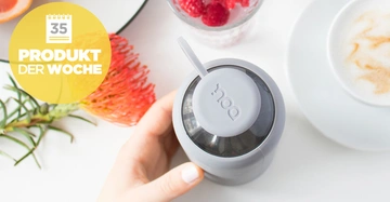 Hand hält grauen Behälter mit Strohhalm auf Tisch neben Kaffee Tasse Blumen und Schale mit Himbeeren. Gelber Kreis mit Text 35 PRODUKT DER WOCHE