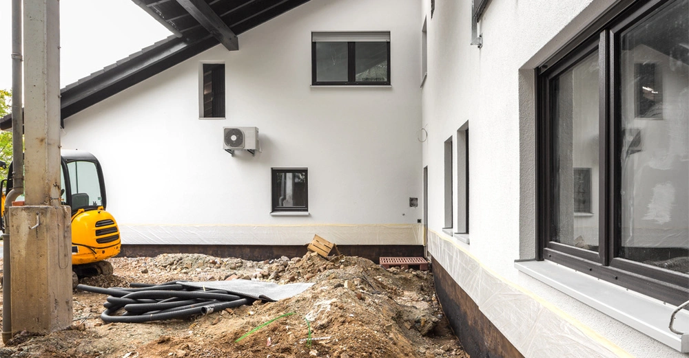 älteres, bewohntes Einfamilienhaus mit aufgegrabenen Kellermauern zur nachträglichen Abdichtung oder Einbringung einer Drainage. Bungalow ist weiß verputzt, Wetter bewölkt