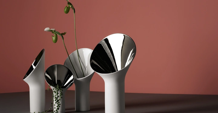 Galerie-Bild 6: Weiße Vasen mit schräg geschnittenen Rändern halten grüne Pflanzen in einem dunklen Raum vor einer rosafarbenen Wand.