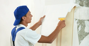 Ein Arbeiter in blauem Overall und Mütze zieht Tapete von einer Wand ab in einem renovierungsbedürftigen Raum.