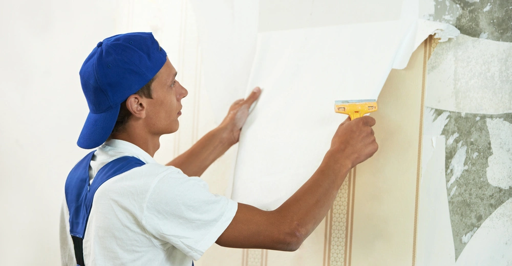 Ein Arbeiter in blauem Overall und Mütze zieht Tapete von einer Wand ab in einem renovierungsbedürftigen Raum.