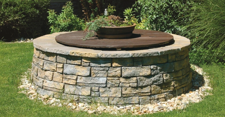 Galerie-Bild 6: Rundbrunnen mit Natursteinmauer und hölzerner Abdeckung auf der eine bepflanzte Schale steht umgeben von gepflegtem Rasen und üppigen grünen Sträuchern in einem Garten.