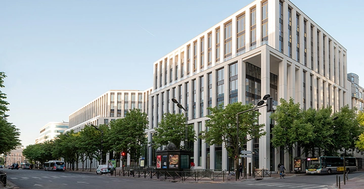 2 Gebäude Komplex aus weißem Sichtbeton mit viel Fensterflächen, im Vordergrund Straße mit Verkehr und Alleebäumen, blauer Himmel