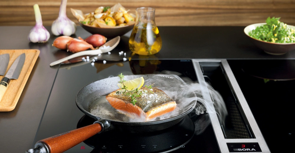 Eine Pfanne brät ein Stück Lachs auf einem Induktionskochfeld. Im Hintergrund stehen Knoblauch, Zwiebeln, Kartoffeln, Öl und eine Schüssel mit grünen Bohnen auf der Küchenarbeitsplatte. Text: BORA.