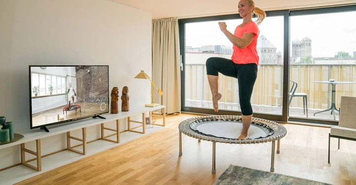 Eine Frau trainiert auf einem Trampolin in einem modernen Wohnzimmer mit Holzfußboden und großen Fenstern, während ein Fitnessvideo auf dem Fernseher läuft