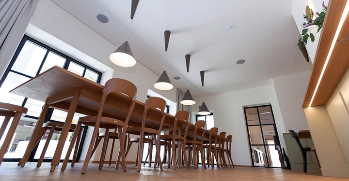 Galerie-Bild 4: Mehrere Holzstühle stehen um einen großen rechteckigen Holztisch. Hängelampen hängen von der Decke. Der Raum ist modern eingerichtet mit großen Fenstern und weißen Wänden.