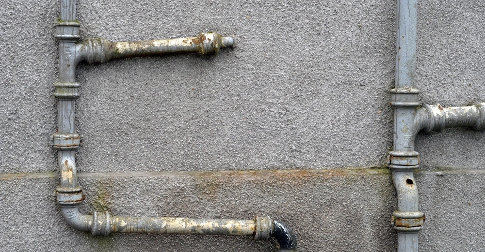 Alte Metallrohre verlaufen horizontal und vertikal entlang einer grauen Betonwand einige Rohre sind rostig und haben Gebrauchsspuren.