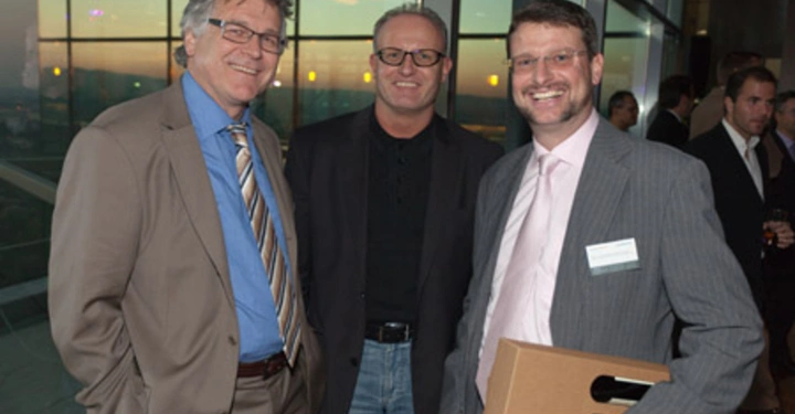 Galerie-Bild 55: Drei Männer stehen lächelnd zusammen und posieren für ein Foto in einem modernen Gebäude mit Glaswänden im Hintergrund Ein Mann hält eine kleine Schachtel Die Umgebung vermittelt ein professionelles Event
