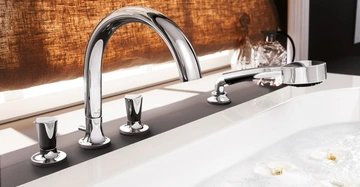 Ein verchromter Wasserhahn füllt eine Badewanne, die von schwimmenden weißen Blüten und Schaum umgeben ist; dahinter ist ein brauner Vorhang.