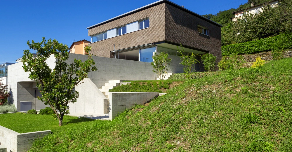Modernes zweigeschossiges Haus steht auf einer grünen Anhöhe vor blauem Himmel Das Haus hat große Fenster und eine Ziegel- sowie Betonfassade und ist von Bäumen und Sträuchern umgeben