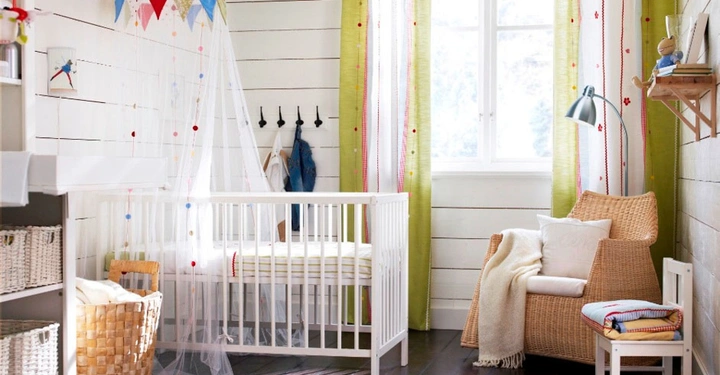 Galerie-Bild 2: Babybett gestanden unter einem bunten Betthimmel neben einem Weidenstuhl in einem hellen Zimmer mit grünen Vorhängen Körben Regalen und einem Fenster im Hintergrund