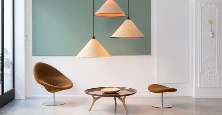 Zwei braune Stühle umgeben einen niedrigen runden Tisch vor einer grünen Wand drei hängende Lampen leuchten darüber in einem minimalistisch eingerichteten Raum mit großen Fenstern links.