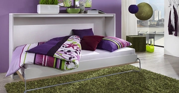 Ein ausklappbares Bett mit bunter Bettwäsche ist ausgeklappt im Wohnzimmer mit lila Wänden und grünen Dekorationen umliegender Möbel zu sehen. Große Fenster bieten Blick auf Stadtlandschaft.