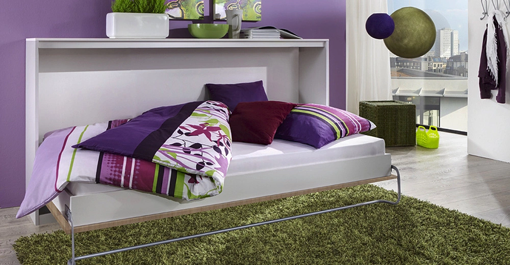 Ein ausklappbares Bett mit bunter Bettwäsche ist ausgeklappt im Wohnzimmer mit lila Wänden und grünen Dekorationen umliegender Möbel zu sehen. Große Fenster bieten Blick auf Stadtlandschaft.