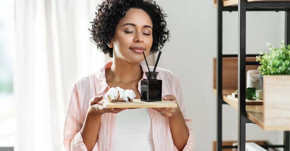 junge Frau mit lockigem schwarzem Haar steht in ihrem modernen Wohnzimmer und schnuppert begeistert an Duftstäbchen. 