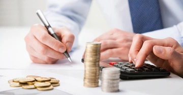 Hände stapeln Münzen und nutzen einen Taschenrechner auf einem Schreibtisch mit Diagrammen und Papieren in einem Büro.