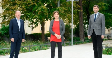 Drei Personen stehen lächelnd nebeneinander die Umgebung ist ein gepflasterter Weg mit Bänken und grünen Bäumen im Freien die Menschen sind formell aber unterschiedlich gekleidet.