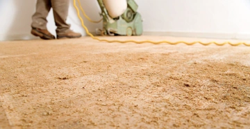 Person schleift den Boden mit einer Schleifmaschine in einem Raum. Der Boden ist mit Sägemehl bedeckt und das gelbe Kabel der Maschine ist zu sehen.