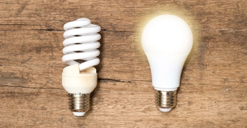 Zwei Glühbirnen liegen auf einer Holzoberfläche. Die linke Energiesparlampe ist ausgeschaltet. Die rechte LED-Lampe leuchtet.