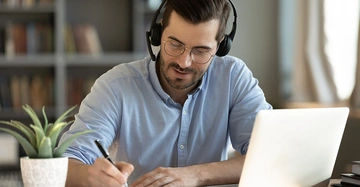 Mann trägt Kopfhörer und Brille schreibt auf Papier vor einem Laptop in einem Zuhause mit Bücherregalen im Hintergrund und einer kleinen Pflanze auf dem Schreibtisch