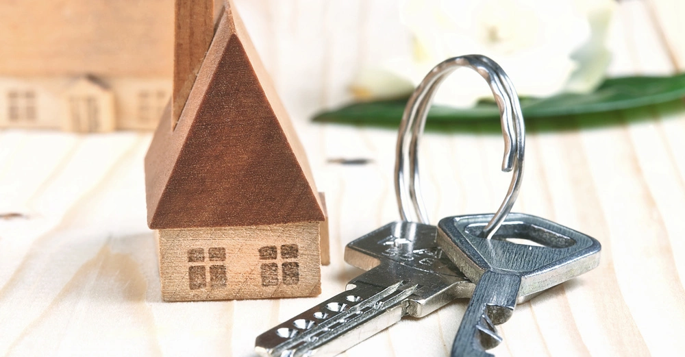 Miniaturhaus aus Holz stehend neben zwei Metall-Schlüsseln auf einem Holztisch. Im Hintergrund unscharfe grüne Blätter und weiße Blüten.