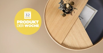 Runder Holztisch beherbergt eine Vase mit Trockenblumen und Bücher daneben gelb-weißer Kreis mit der Aufschrift 32 PRODUKT DER WOCHE auf einem beigen Hintergrund.