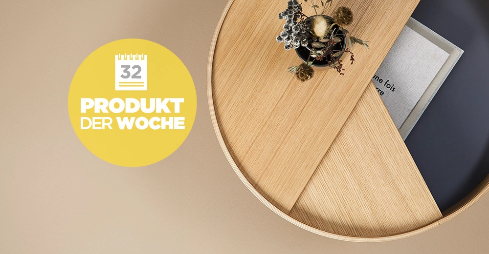Runder Holztisch beherbergt eine Vase mit Trockenblumen und Bücher daneben gelb-weißer Kreis mit der Aufschrift 32 PRODUKT DER WOCHE auf einem beigen Hintergrund.
