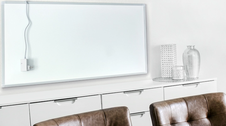 Ein weißes Infrafrotpaneel in einem minimalistischen Wohnraum über einem weißen Regal und einem Tisch mit braunen Ledersesseln.