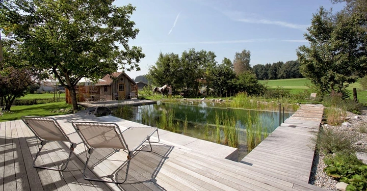Zwei Liegestühle stehen auf einer Holzterrasse ruhig am Rand eines kleinen Teiches in einem grünen Garten mit Bäumen und einem kleinen Holzhaus im Hintergrund.