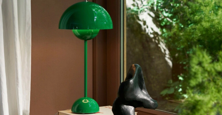 Eine grüne Tischlampe steht auf einem Holzstapel neben einer schwarzen Skulptur vor einem Fenster mit Blick ins Grüne und einem weißen Vorhang links.