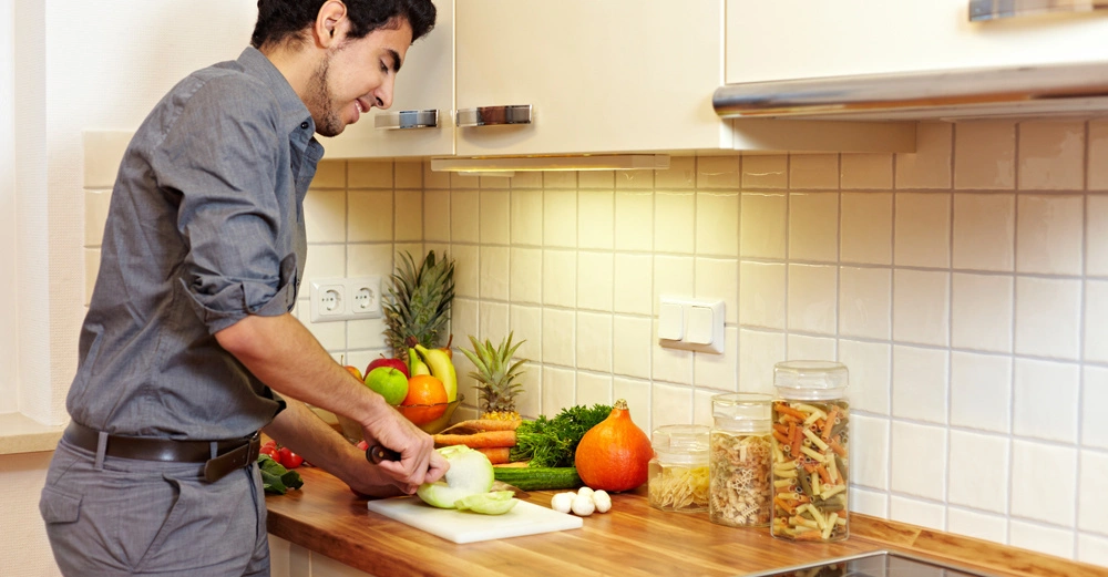 Ein Mann schneidet Gemüse auf einem Schneidebrett in einer modernen Küche mit weißen Fliesen und Holzarbeitsplatte. Daneben stehen weitere Gemüse und Gläser mit Nudeln und Reis.