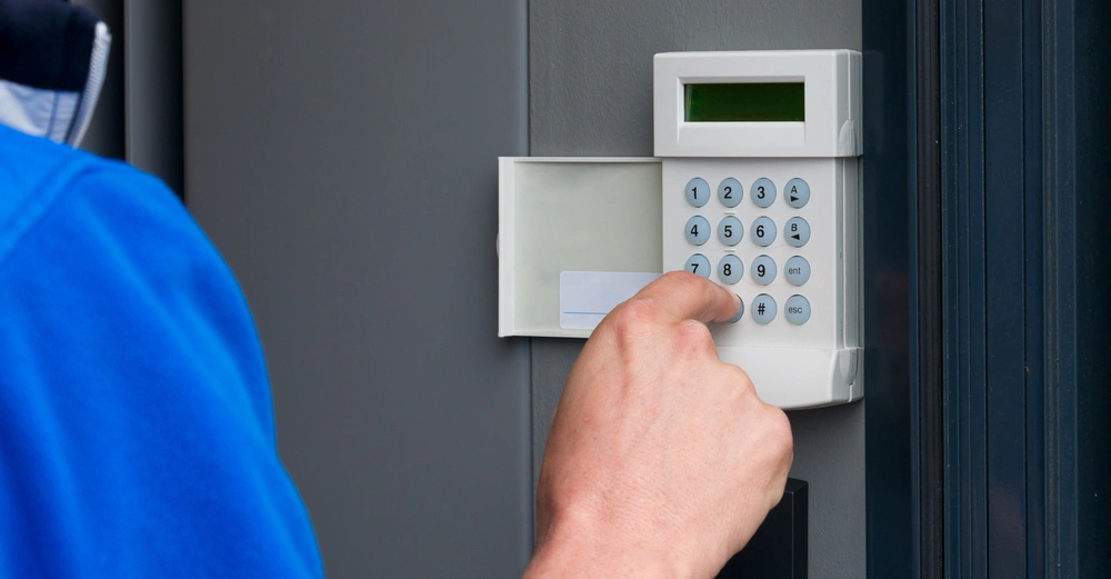 Eine Hand drückt Tasten auf einem weißen Tastenfeld einer Alarmanlage an einer grauen Wand in der Nähe einer Tür