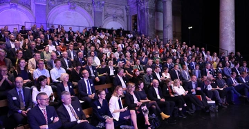 Eine Vielzahl von Personen sitzt in formeller Kleidung und hört zu in einem großzügigen, klassisch gestalteten Saal mit Säulen und beleuchteten Wände.