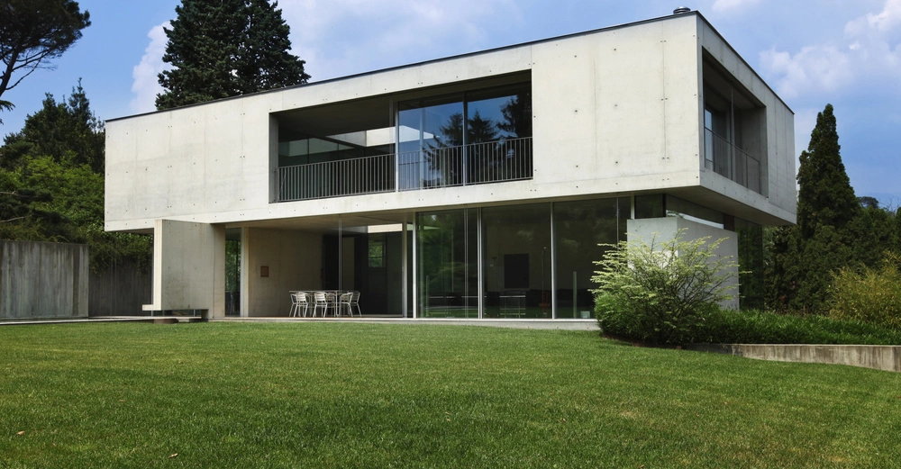 Ein modernes Betonhaus steht in einem gepflegten Garten mit großem Rasenbereich und umgeben von hohen Bäumen. Auf dem Balkon des Hauses steht eine Sitzgruppe.