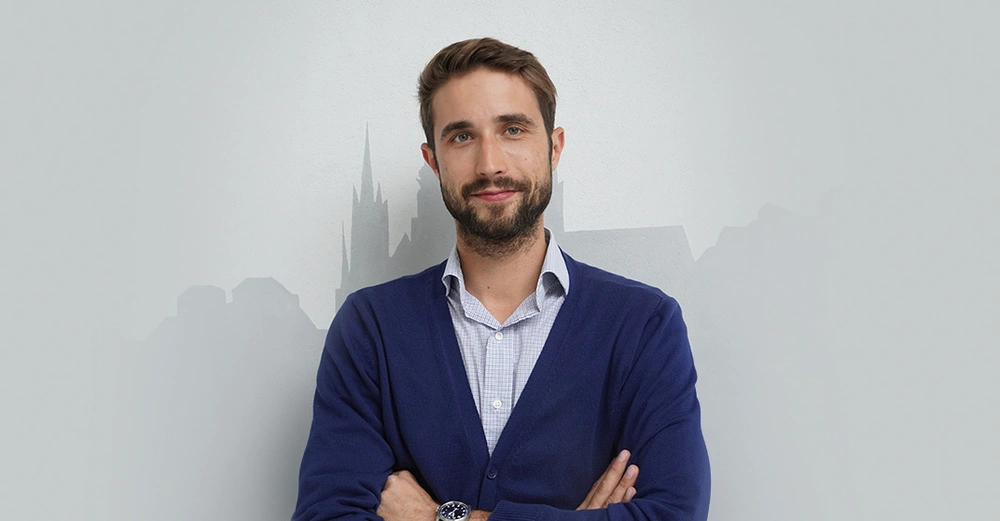 Portrait von R. Bergsleithner vor weißer Wand, verschränkte Arme, in weißem Hemd und blauer Weste, freundlich lächelnd