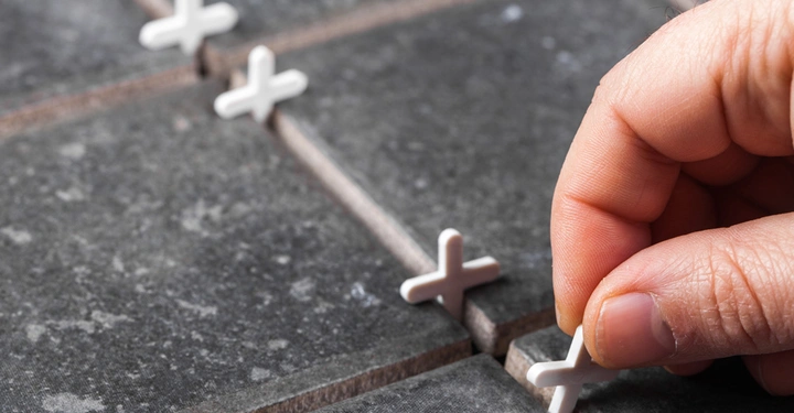 Eine Hand platziert weiße Fliesenkreuze zwischen dunklen Fliesen auf einer Oberfläche.