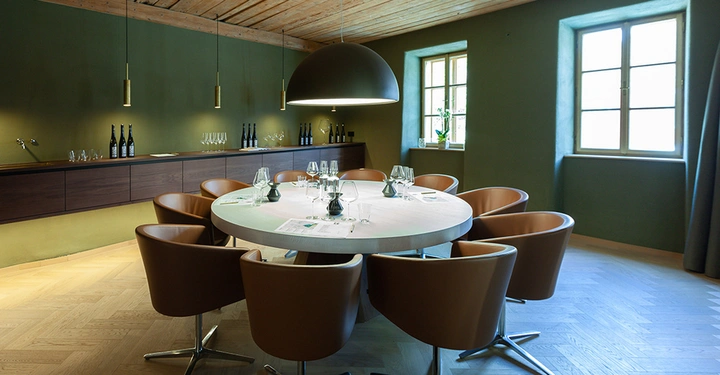 Galerie-Bild 6: Großer runder Tisch mit Lederstühlen, gedeckt mit Weingläsern und Tischdekor, in einem gemütlichen Raum mit grünen Wänden, Holzboden und zwei Fenstern; im Hintergrund eine Anrichte mit Weinflaschen und Gläsern.