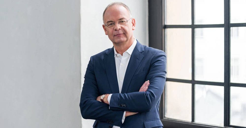 Ein Mann steht mit verschränkten Armen in einem dunkelblauen Anzug in einem hell beleuchteten Raum neben einem großen Fenster mit schwarzen Gittern.