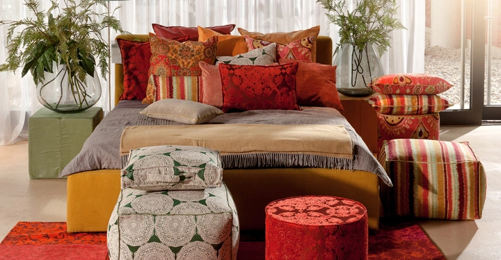 Bett mit bunten Kissen in verschiedenen Mustern Steht in einem hellen Raum Pflanzen in Vasen auf beiden Seiten Rote und grüne Hocker und Poufs auf Teppich ergänzt die Szenerie