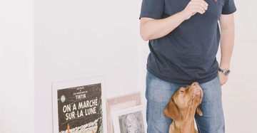 Ein Mann steht neben einem Hund in einem Raum mit hellen Wänden und Kunstwerken. Das Poster zeigt den Text: Hergé Les Aventures de Tintin ON A MARCHÉ SUR LA LUNE.