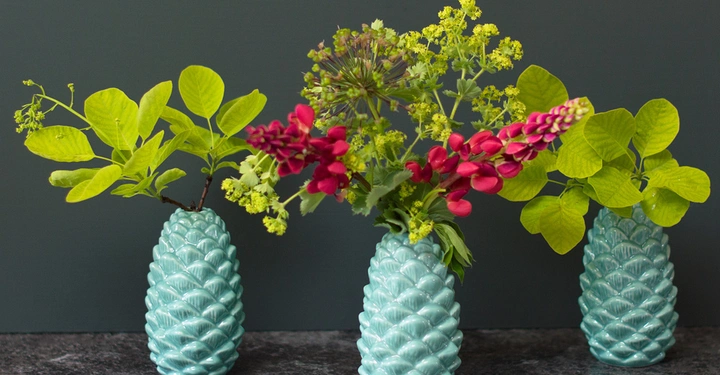 Galerie-Bild 3: Drei türkisfarbene Vasen mit verschiedenen Blumensträußen aus grünen Blättern und roten Blüten stehen auf einer dunklen Oberfläche vor einem dunklen Hintergrund.