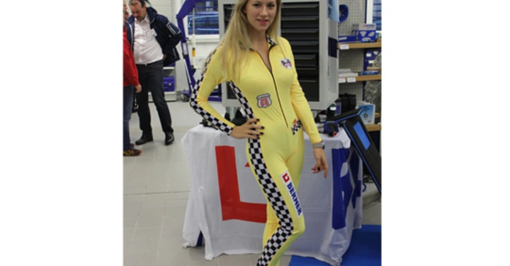 Galerie-Bild 4: Eine Frau in einem gelben Rennanzug posiert in einem Verkaufsraum für Werkzeuge. Schilder im Hintergrund zeigen Beschriftungen wie Elektro-Werkzeug. Weitere Personen sind im Hintergrund sichtbar.