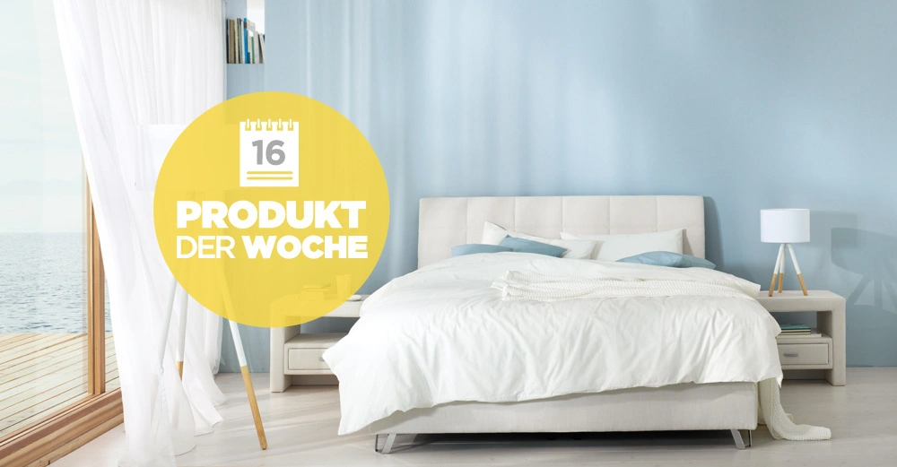 Bett steht im hellen Schlafzimmer mit blauen Wänden und weißen Vorhängen neben einem Fenster mit Meerblick Text auf gelbem Kreis 16 PRODUKT DER WOCHE