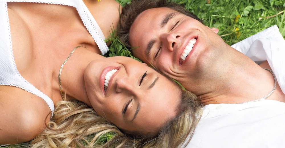 Ein Mann und eine Frau liegen nebeneinander auf dem Gras und lächeln beide mit geschlossenen Augen. Die Frau trägt eine weiße Bluse und eine Halskette mit einer gelben Blume im Haar.