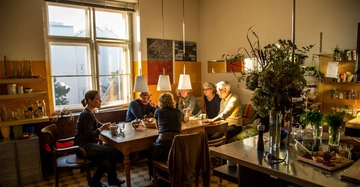 Personen sitzen an einem Holztisch und unterhalten sich in einer hell erleuchteten Küche mit großen Fenstern und einer gemütlichen Atmosphäre. Pflanzen und Küchengeräte umgeben den Raum.