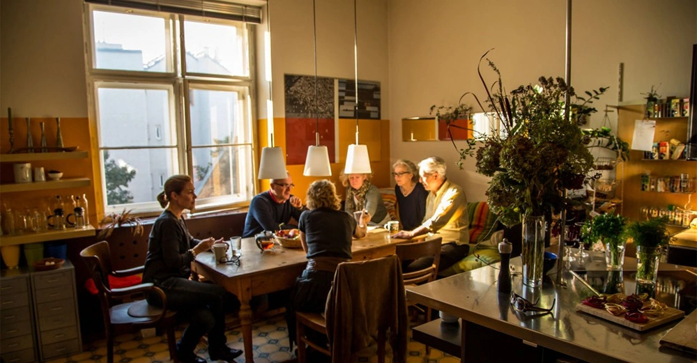 Personen sitzen an einem Holztisch und unterhalten sich in einer hell erleuchteten Küche mit großen Fenstern und einer gemütlichen Atmosphäre. Pflanzen und Küchengeräte umgeben den Raum.