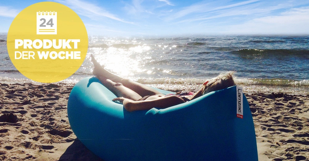 Person liegt auf einem aufblasbaren Liegesack am Strand und sonnt sich; das Meer im Hintergrund. Gelbes Badge mit Text 24 Produkt der Woche in der oberen linken Ecke.