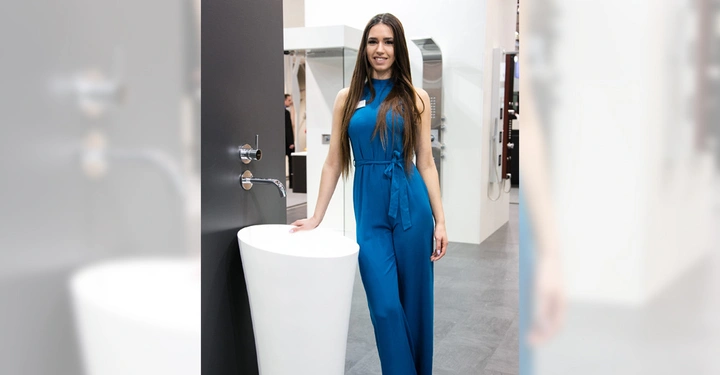 Galerie-Bild 11: Frau in blauem Overall neben modernem, freistehendem Waschbecken in Ausstellungsbereich für Badezimmerausstattung. Sie lächelt und hält die Hand auf dem Beckenrand in einem hell beleuchteten Raum