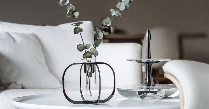 Galerie-Bild 7: Eine kugelförmige Vase hält Eukalyptuszweige auf einem weißen runden Tisch in einem modernen Wohnzimmer mit weißen Möbeln. Daneben steht eine zweistufige Servierplatte.